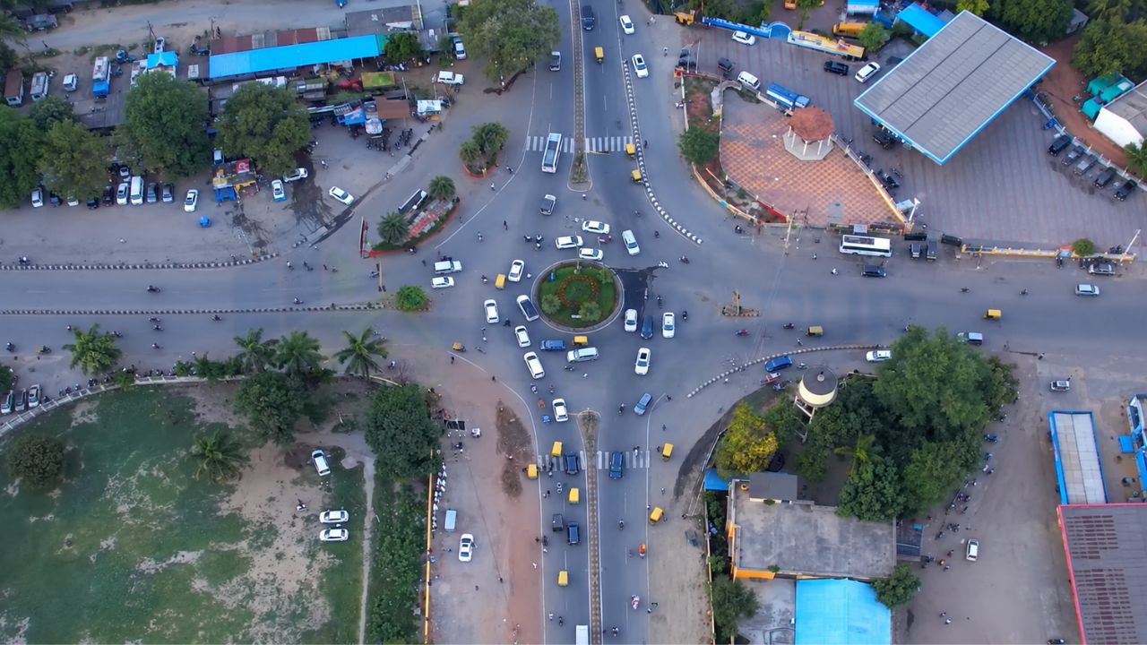 Aerial drone view of a major traffic roundabout in Rudrapur showing road connectivity, commercial surroundings, vehicles, and urban infrastructure.