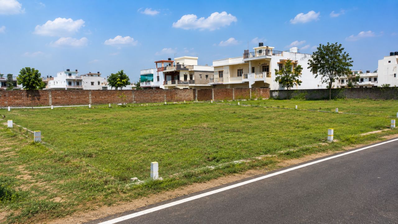 Freehold residential plot in Rudrapur with a developed road, boundary wall, and nearby homes.